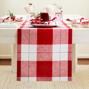 May include: A rectangular table runner featuring a bold red and white buffalo check pattern with thin green accent lines. The fabric has a visible woven texture and hangs over the edge of a white wooden table set with plates and glassware.