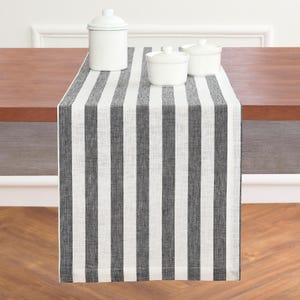 May include: A black and white striped table runner with three white ceramic containers on top. The runner is made of a textured fabric and is draped over a wooden table. The containers have lids and are of varying sizes.