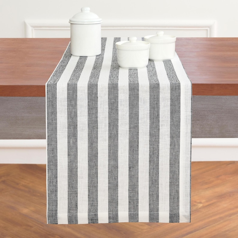 May include: A black and white striped table runner with three white ceramic containers on top. The runner is made of a textured fabric and is draped over a wooden table. The containers have lids and are of varying sizes.