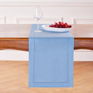 May include: A light blue table runner with a stitched border, displayed on a wooden table. A white serving dish filled with red grapes and a clear wine glass are on the table.