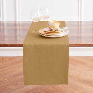 May include: A mustard-colored table runner is draped over a wooden table. Two wine glasses, a stack of plates, bowls, and silverware are arranged on the runner. The runner is made of a textured fabric.