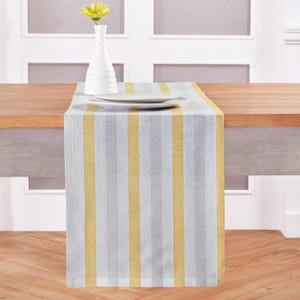 May include: A striped table runner in yellow, gray, and white, placed on a wooden table. A white vase with a yellow flower and two white plates are on top of the runner. The runner's design features vertical stripes.