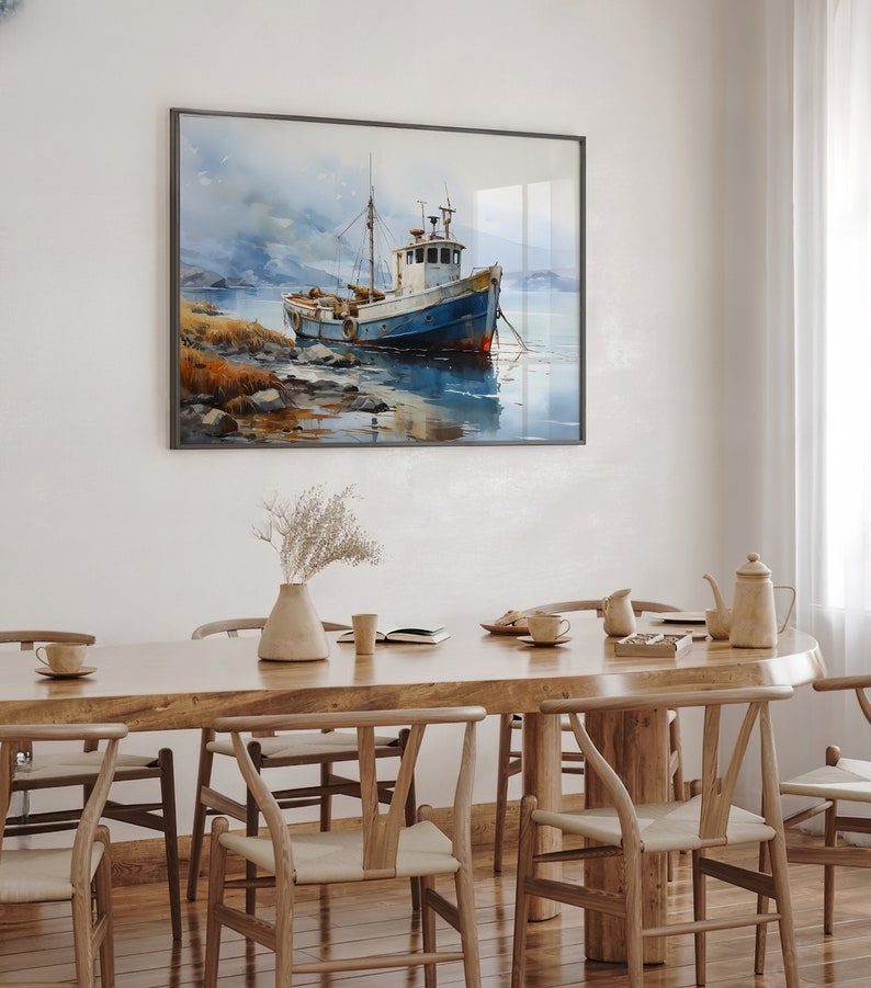 May include: A watercolour painting of a blue and white fishing boat moored in a harbour. The boat is surrounded by water and a rocky shore. The painting is framed in a black frame and hangs on a white wall above a wooden dining table with chairs.
