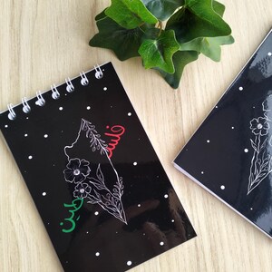 Peut inclure: Un carnet à spirales noir avec un contour blanc de la Palestine avec des accents rouges et verts et le mot "Palestine" écrit en arabe. Le carnet a des points blancs sur un fond noir.