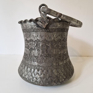 May include: Antique silver-colored metal bucket with a handle. The bucket has an embossed floral design around the body and a textured diamond pattern at the base. The rim is scalloped.