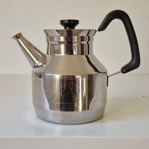 May include: A stainless steel teapot with a black handle and lid. The teapot has a spout and a curved handle. The teapot is sitting on a white surface.