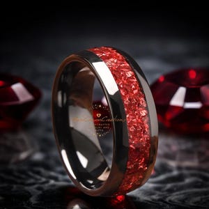Puede incluir: Un anillo de carburo de tungsteno negro con una incrustación roja. La incrustación está hecha de vidrio rojo triturado y está engastada en el anillo.