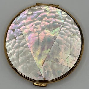 May include: A round, vintage compact with a gold-tone metal frame. The top features a mosaic design of iridescent, cracked mother-of-pearl pieces, reflecting hues of pink, green, and yellow. The compact is closed, showing its complete circular form.