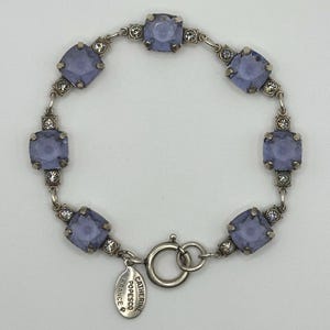 May include: A silver-toned bracelet featuring six square-cut lavender crystals alternating with small clear rhinestones. The piece includes a circular clasp and an oval metal tag engraved with the text CATHERINE POPESCO FRANCE.
