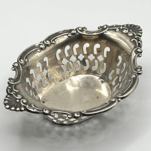 May include: An oval silver-toned metal basket featuring an intricate openwork lattice pattern on the sides. The rim is decorated with embossed scrollwork and shell-like motifs at the ends, creating a polished, reflective surface throughout.