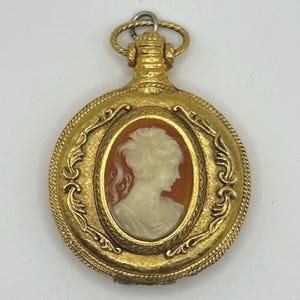 May include: A gold-toned pocket watch pendant with an oval cameo of a woman in profile. The cameo is set against an orange background and framed by ornate gold filigree. The watch has a decorative loop at the top.