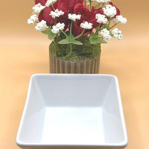 May include: A square, white ceramic bowl with a glossy finish. Behind the bowl is a decorative arrangement of red roses and white flowers in a brown, textured pot. The background is a solid, light orange color.