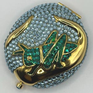 May include: An oval compact with a gold-tone frame, covered in light blue rhinestones. The design features a gold-tone dragon and green rhinestones. A small, blue jewel is on the bottom. The compact is a decorative item.