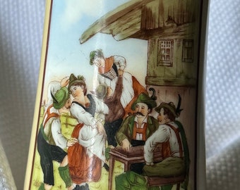May include: A vintage ceramic beer stein with a pewter lid. The stein features a colorful illustration of people in traditional German clothing dancing and playing music. The text "Schopflattler" is visible at the bottom of the stein.