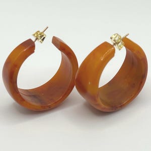 Peut inclure: Une paire de boucles d'oreilles créoles fabriquées dans une résine translucide de couleur ambre avec un motif marbré. Chaque boucle présente une forme en C épaisse et est dotée d'une tige en métal doré avec un fermoir papillon assorti.