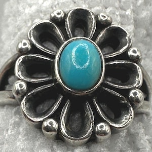 Puede incluir: Anillo de plata con una piedra turquesa en el centro, rodeada por un diseño floral. El anillo tiene un acabado oxidado detallado, con pequeñas cuentas de plata que acentúan los pétalos. La piedra turquesa es de forma ovalada.
