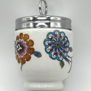 Puede incluir: Un cocedor de huevos de cerámica blanca con una tapa de metal cromado pulido que tiene un asa circular. El cuerpo está decorado con coloridos motivos florales en tonos naranja, azul cerceta, morado y verde, mostrando flores y hojas.