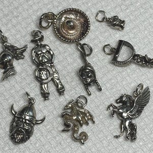 Op de afbeelding: Een verzameling van tien zilveren bedelarmbanden met verschillende ontwerpen, waaronder een cowboy, een draak, een gevleugeld paard, een sombrero, een viking en een indiaanse hoofdtooi.