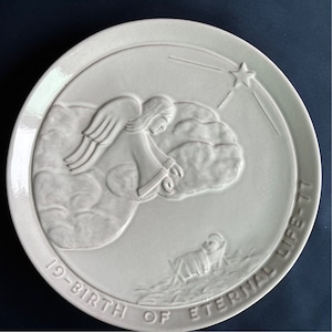 May include: White ceramic plate with a raised design of an angel with wings, a star, and a baby in a manger. The plate has the text "19-Birth of Eternal Life" around the edge.
