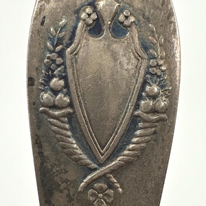 May include: Close-up of a silver spoon with an ornate design. The spoon features a shield with a floral wreath and a small flower at the bottom.