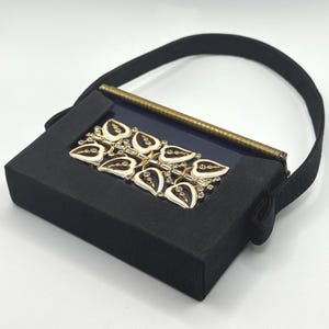 May include: A black rectangular handbag with a black handle. The front features a gold-toned leaf design with white enamel and small clear rhinestones. A gold-toned pen is inside the bag.