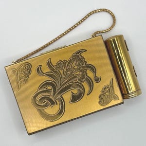 May include: A vintage gold-toned compact with a matching lipstick holder. The compact features an engraved floral design and a delicate chain handle. The lipstick holder is cylindrical and also gold-toned.
