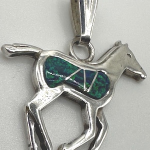 Puede incluir: Colgante de plata con forma de caballo. El caballo tiene un diseño de incrustaciones verdes y azules. El colgante tiene un lazo en la parte superior para una cadena. El caballo está en movimiento.