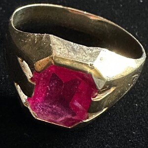 May include: A gold ring featuring a large, faceted, red gemstone. The ring has a simple, geometric design.
