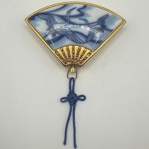 以下が含まれることがあります： 金色のフレームが付いた扇形のブローチ。扇面は、青と白の鶴のデザインが施された磁器製です。扇の基部には、小さなオレンジ色のクリスタルが飾られています。装飾的な結び目の付いた青いコードが下部にぶら下がっています。