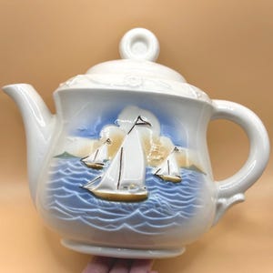 May include: White ceramic teapot with a blue and white seascape design featuring three sailboats. The teapot has a lid with a knob on top and a handle on the side.