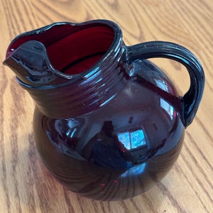 Puede incluir: Una jarra de vidrio de color morado oscuro con cuerpo redondeado y asa. La jarra tiene un acabado liso y brillante.