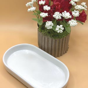 Puede incluir: Una bandeja ovalada de cerámica blanca se encuentra frente a un arreglo floral decorativo. Las flores son una mezcla de rosas rojas oscuras y pequeños racimos blancos, dispuestas en una maceta gris texturizada. El fondo es de color beige claro.