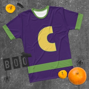 May include: Purple t-shirt with a green and yellow stripe across the bottom. The shirt has a large yellow letter "C" on the front. The shirt is on a grey background with a few small pumpkins and bats.