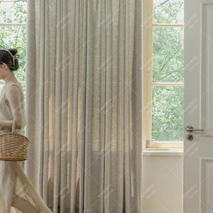 May include: Beige linen curtains with vertical pleats. The curtains are hanging in front of a window with natural light. A person is walking by with a woven basket. The curtains are a neutral color, suitable for various interior design styles.