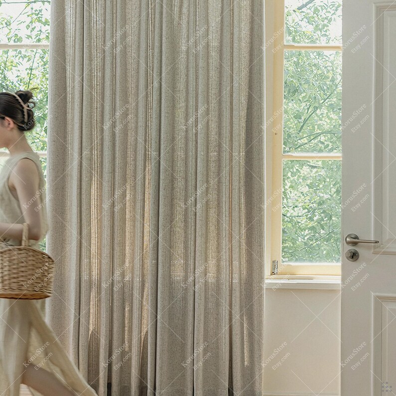 May include: Beige linen curtains with vertical pleats. The curtains are hanging in front of a window with natural light. A person is walking by with a woven basket. The curtains are a neutral color, suitable for various interior design styles.