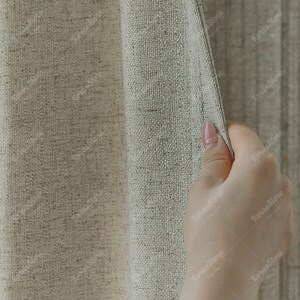 May include: Close-up of a beige linen curtain. The fabric has a textured weave and is being held open by a hand, revealing a glimpse of a similar curtain behind it. The overall color palette is neutral, with shades of beige and tan.