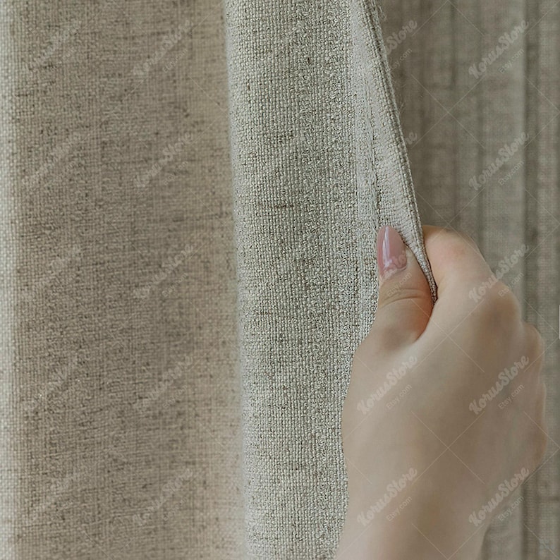 May include: Close-up of a beige linen curtain. The fabric has a textured weave and is being held open by a hand, revealing a glimpse of a similar curtain behind it. The overall color palette is neutral, with shades of beige and tan.