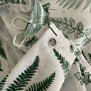 May include: Close-up of a white shower curtain with a green fern pattern. The curtain has metal grommets for hanging.