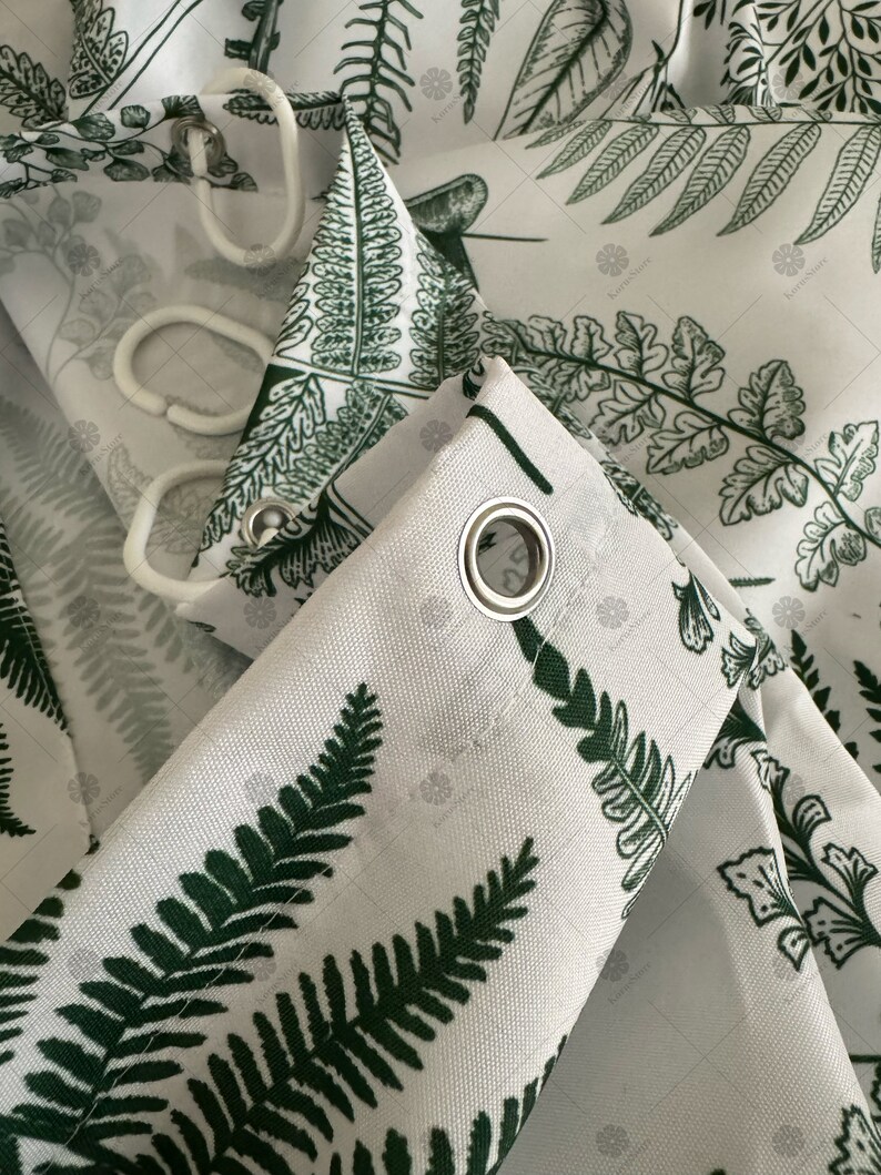 May include: Close-up of a white shower curtain with a green fern pattern. The curtain has metal grommets for hanging.