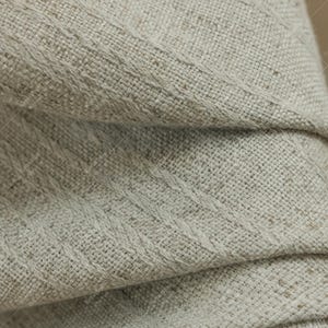 May include: Close-up of a folded, textured fabric in a neutral beige color. The fabric has a woven appearance with diagonal stitched details. The folds create shadows, highlighting the texture and depth of the material.