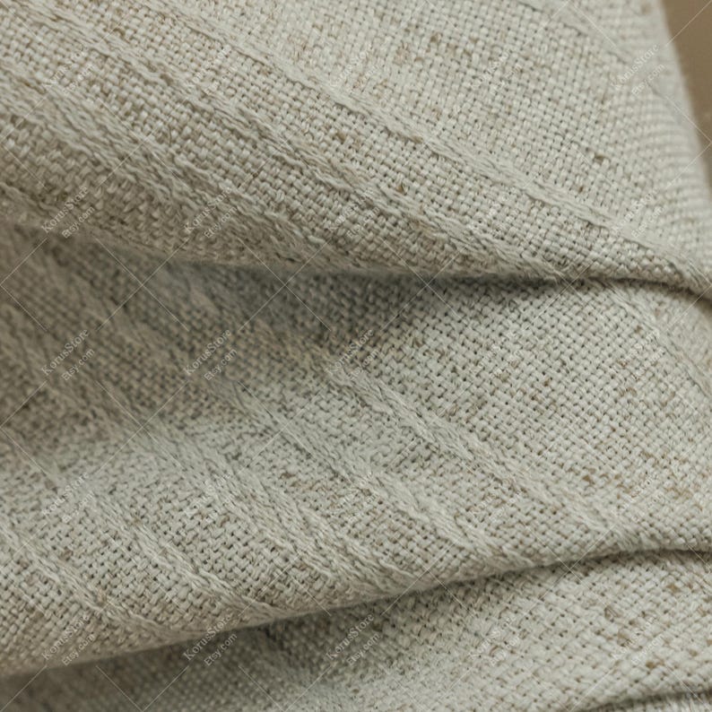 May include: Close-up of a folded, textured fabric in a neutral beige color. The fabric has a woven appearance with diagonal stitched details. The folds create shadows, highlighting the texture and depth of the material.