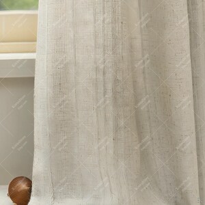 May include: Off-white linen curtains with a textured weave. The sheer panels filter light, creating a soft, diffused glow. The curtains are hanging near a window, adding a touch of elegance to the room. The fabric has a natural, organic look.