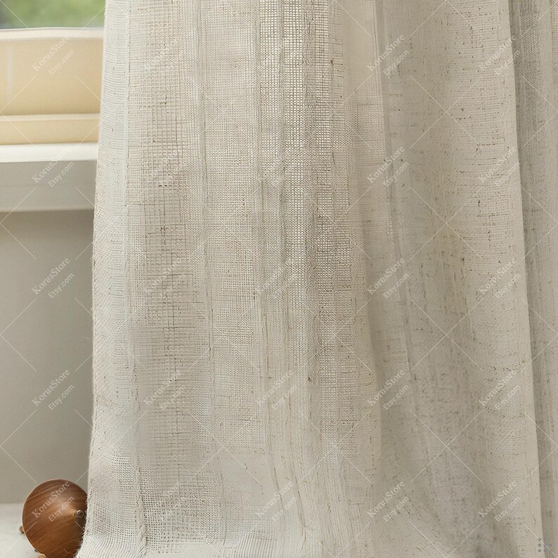 May include: Off-white linen curtains with a textured weave. The sheer panels filter light, creating a soft, diffused glow. The curtains are hanging near a window, adding a touch of elegance to the room. The fabric has a natural, organic look.