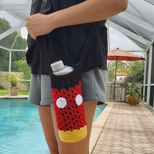 May include: A crocheted water bottle holder with a Mickey Mouse design. The holder is red, black, white, and yellow. It is being worn by a person as a crossbody bag.