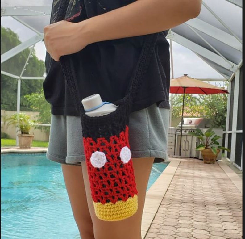 May include: A crocheted water bottle holder with a Mickey Mouse design. The holder is red, black, white, and yellow. It is being worn by a person as a crossbody bag.