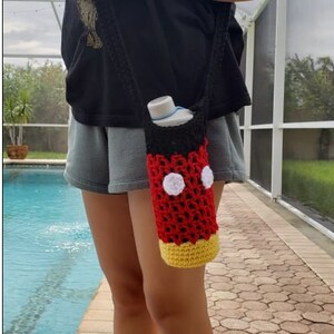 May include: A red, black, and yellow crocheted water bottle holder with white circles. The holder is shaped like Mickey Mouse's head and is being worn as a crossbody bag.
