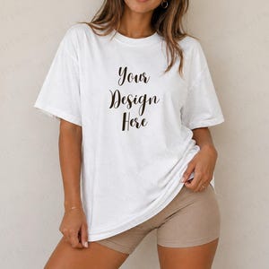 Może przedstawiać: Biała koszulka z napisem "Your Design Here" czcionką w kolorze czarnym. Koszulka jest w rozmiarze oversize i noszona z beżowymi szortami. Modelka ma na sobie złotą bransoletkę.