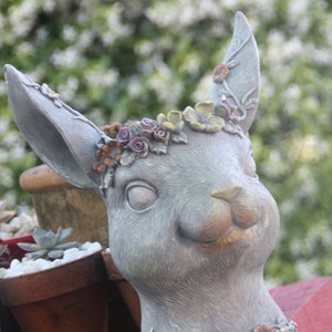 May include: A grey ceramic rabbit planter with a floral crown made of colourful plastic flowers. The planter has a detailed face with a small nose and closed eyes.