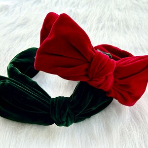 Puede incluir: Dos diademas de terciopelo, una roja y otra verde oscuro, cada una con un gran lazo. Las diademas se muestran sobre una superficie blanca y esponjosa, creando un contraste llamativo de colores y texturas.