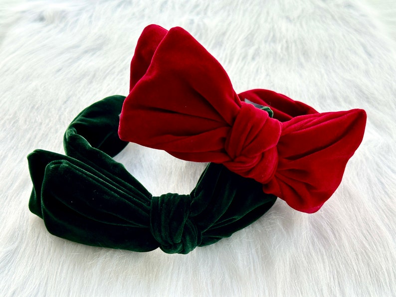 Puede incluir: Dos diademas de terciopelo, una roja y otra verde oscuro, cada una con un gran lazo. Las diademas se muestran sobre una superficie blanca y esponjosa, creando un contraste llamativo de colores y texturas.